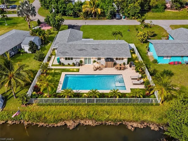 Aerial perspective of a waterfront property featuring a rectangular in-ground swimming pool, expansive paved patio, fenced perimeter, and mature landscaping