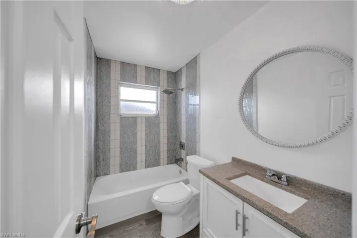 Bathroom featuring a vanity with a stone-finish countertop, oval wall-mounted mirror, wood-finish flooring, and a tub with vertical tile surround