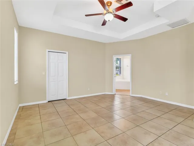 Spacious room featuring neutral-toned tile flooring, light beige wall paint, a tray ceiling, and a ceiling fan with light fixture
