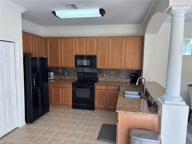 Kitchen with wood finish cabinetry, black appliances, ornate columns, dark stone counters, and a peninsula