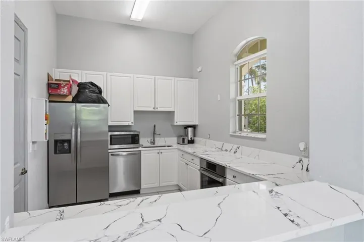 Kitchen located in side community room of clubhouse.  Kitchen featuring white cabinetry, stainless steel appliances, white countertops with dark veining, and an arched window