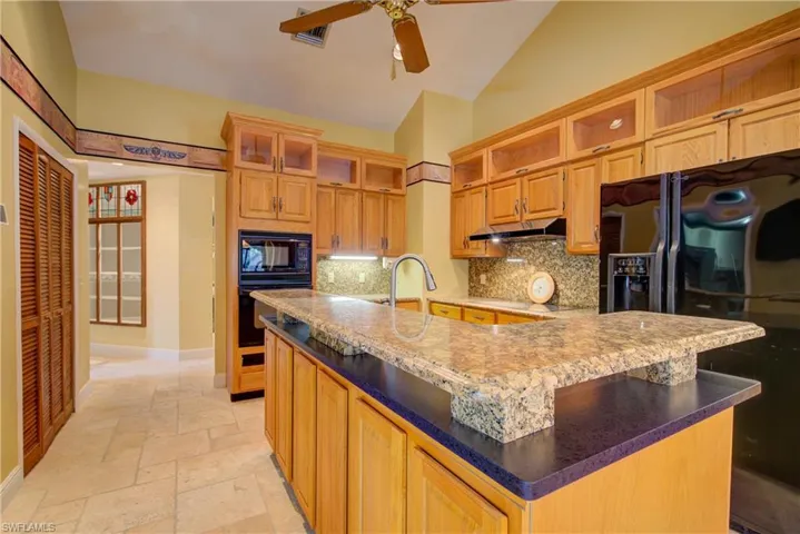 Kitchen featuring black appliances, backsplash, a kitchen island with sink, stone tile floors, and ceiling fan