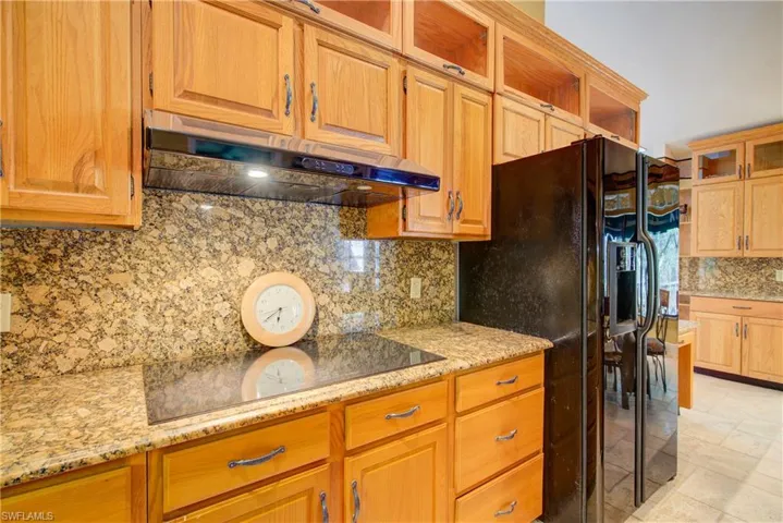 Kitchen with glass insert cabinets, tasteful backsplash, black appliances, under cabinet range hood, and light stone counters
