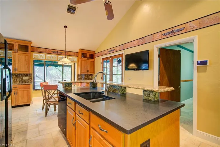 Kitchen featuring brown cabinets, stone tile flooring, a kitchen island with sink, glass insert cabinets, and high vaulted ceiling