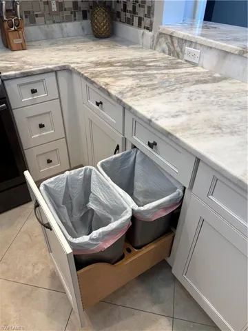 White cabinetry with dark hardware, stone countertops with a backsplash, and a built-in pull-out waste disposal system