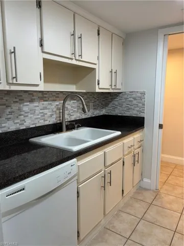 Kitchen featuring dark countertops, dishwashing machine, light tile patterned floors, tasteful backsplash, and white cabinets