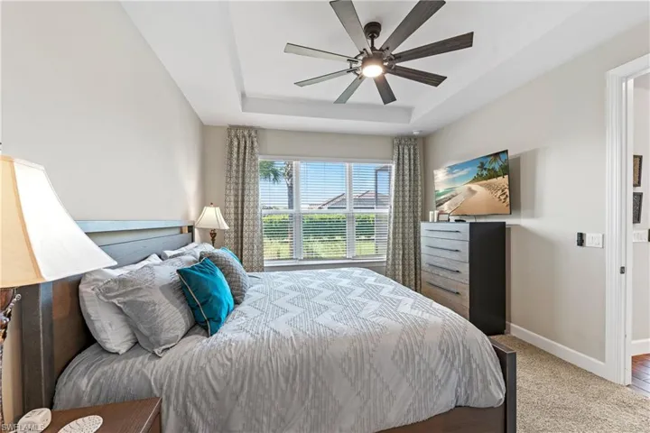 Spacious sleeping area featuring a tray ceiling, integrated ceiling fan with lighting, ample natural light from a window with blinds and curtains, neutral wall tones, and carpet flooring
