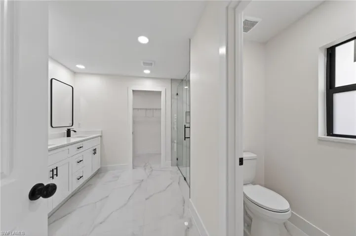 Contemporary bathroom featuring large format marble-look tile flooring, a white vanity with matte black hardware, a frameless glass shower enclosure, recessed lighting, and a separate water closet
