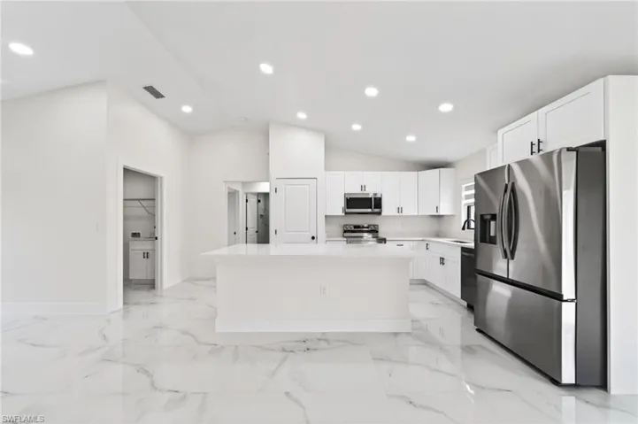 Open-concept kitchen featuring white shaker cabinetry with black hardware, a center island with countertop seating, white marble-look tile flooring, and vaulted ceilings with recessed lighting