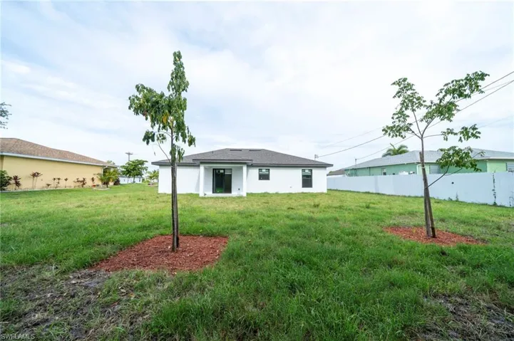 Expansive backyard featuring a manicured lawn, two young trees with mulch beds, a white privacy fence along one boundary, and a modern single-story residence with white stucco exterior, dark roof shingles, and large windows