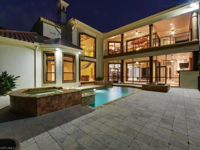 Back of house at night featuring a pool with connected hot tub, a balcony, stucco siding, and a patio