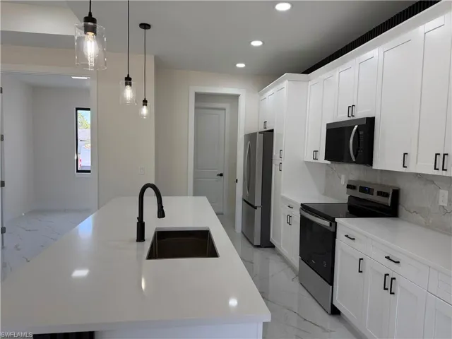 Kitchen with a kitchen island with sink, stainless steel appliances, light marble finish flooring, white cabinetry, and hanging light fixtures