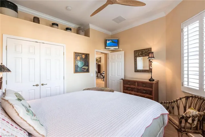 Double-door closet with overhead display ledge, ceiling fan with natural wood-tone blades, white louvered window shutters, and crown molding