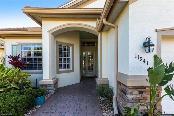 Stucco exterior featuring a stone-veneer foundation, a recessed arched entryway, a brick-paver walkway, and a glass-paneled front door with a transom window