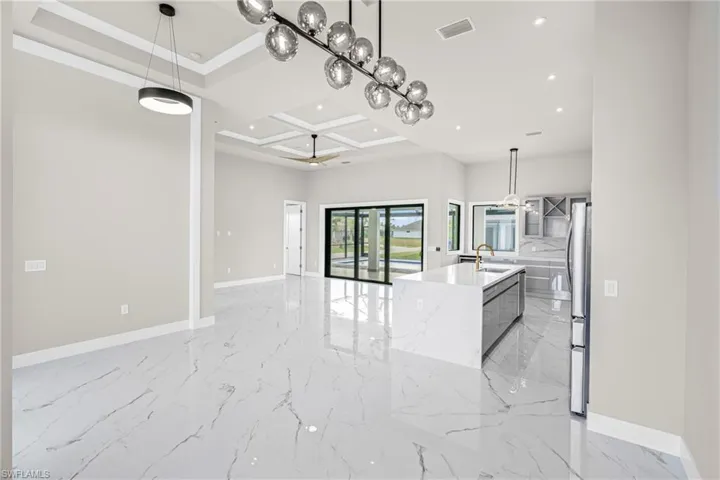 Kitchen with coffered ceiling, decorative light fixtures, a kitchen island with sink, light marble finish flooring, and light stone counters