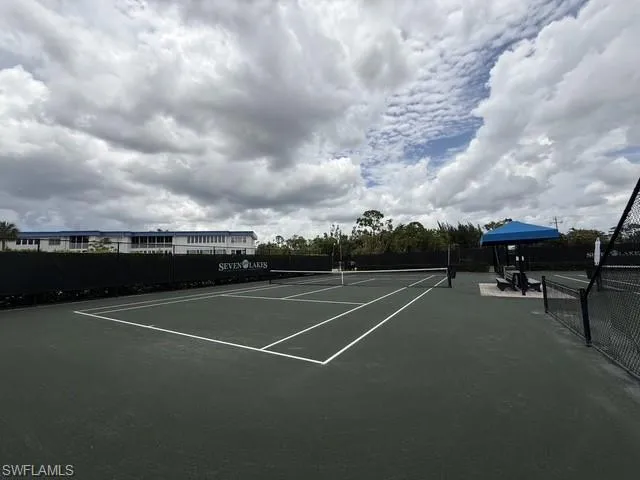Tennis court featuring a dark green playing surface with white lines, black perimeter fencing, a blue shaded seating area, and lush green landscaping