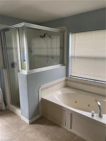 Bathroom featuring a jetted soaking tub, a glass-enclosed shower, light-toned tiled flooring, and a window with blinds