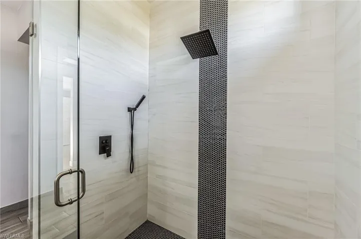 Walk-in shower featuring a frameless glass enclosure, white tile walls with subtle veining, and a vertical accent stripe of small black hexagonal tiles