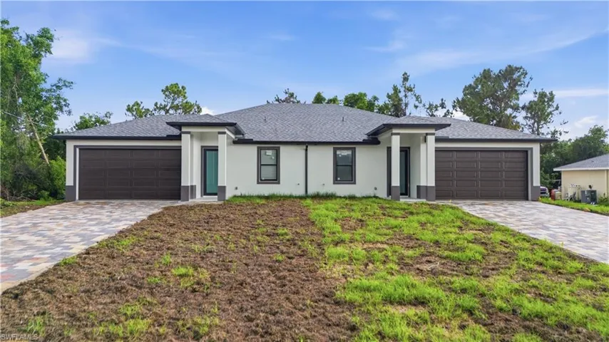 Duplex exterior featuring light-colored stucco, dark trim details, a dark shingle roof, and paver driveways leading to two individual garages with dark paneled doors