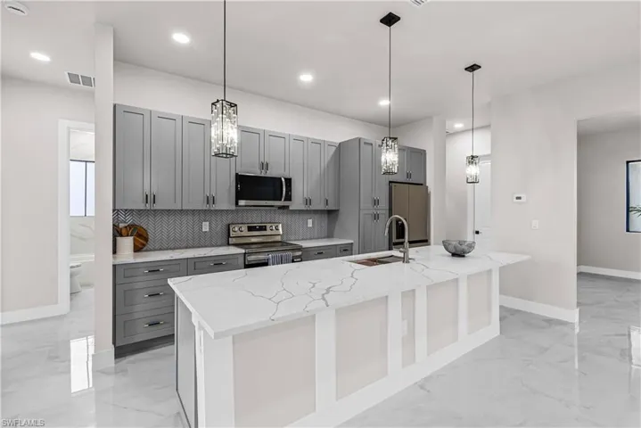 Kitchen with gray cabinetry, stainless steel appliances, pendant lighting, an island with sink, and light stone countertops