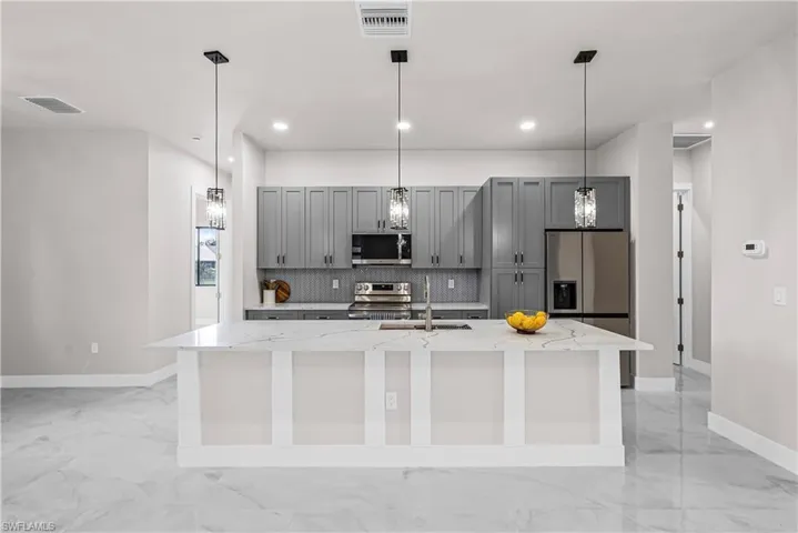 Kitchen with gray cabinets, stainless steel appliances, light stone countertops, tasteful backsplash, and a kitchen island with sink