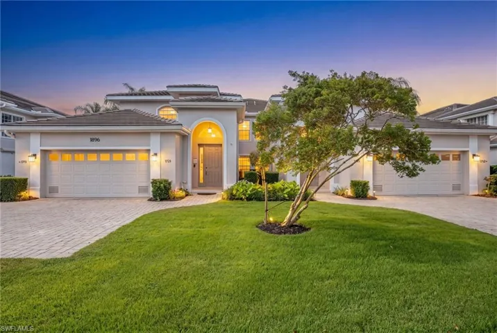 Stucco exterior residence featuring an arched portico entry, two two-car garages with panel doors, paver driveway, and a manicured lawn with mature landscaping