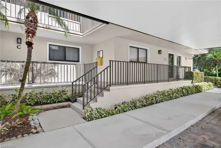 Covered exterior entryway featuring a concrete walkway, elevated stairs with metal railings, and landscaped shrubbery