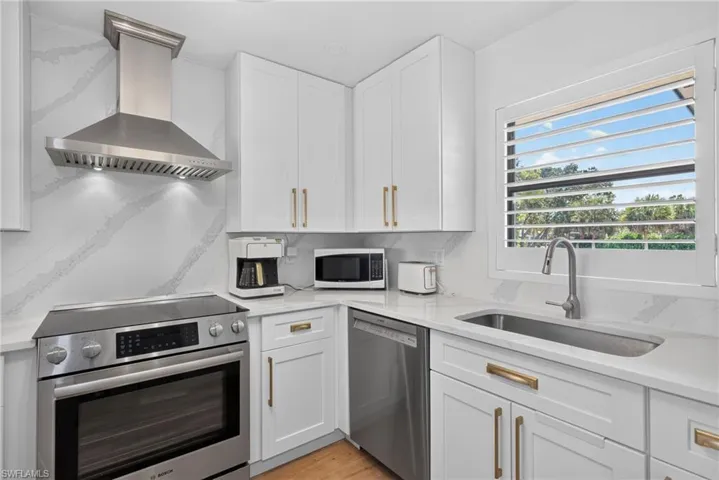 Kitchen featuring white shaker-style cabinetry, quartz countertops with integrated backsplash, stainless steel appliances, a professional-style range hood, and shutters