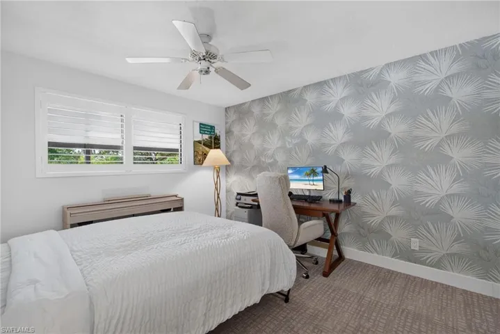 Room featuring a decorative accent wall with palm leaf patterned wallpaper, neutral carpeting, white horizontal shutter window coverings, and a ceiling fan with integrated lighting