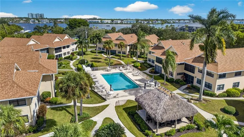 Aerial complex perspective featuring a central swimming pool, a thatched-roof pavilion, paved walkways, mature palm trees, and residential buildings with shingled roofing
