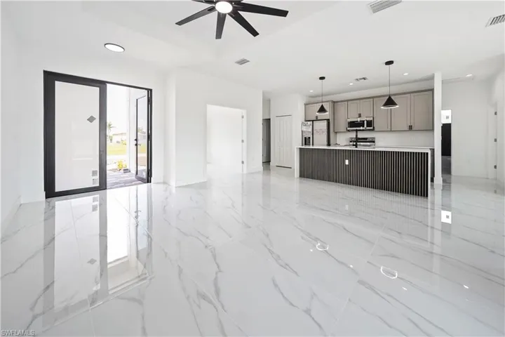 Spacious open-concept living area featuring large format white tile flooring, contemporary kitchen island with ribbed detailing, recessed lighting, modern ceiling fan, and double entry doors with frosted glass panels