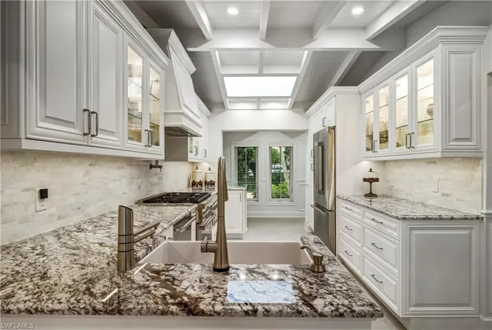 Kitchen with glass insert cabinets, white cabinetry, tasteful backsplash, light stone countertops, and beamed ceiling