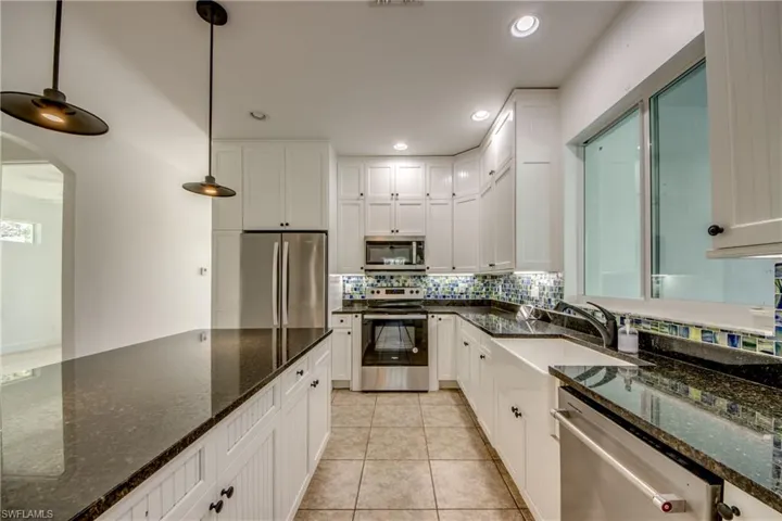 Kitchen featuring dark granite stone counters, stainless steel appliances, light tile flooring, hanging light fixtures, and white cabinetry