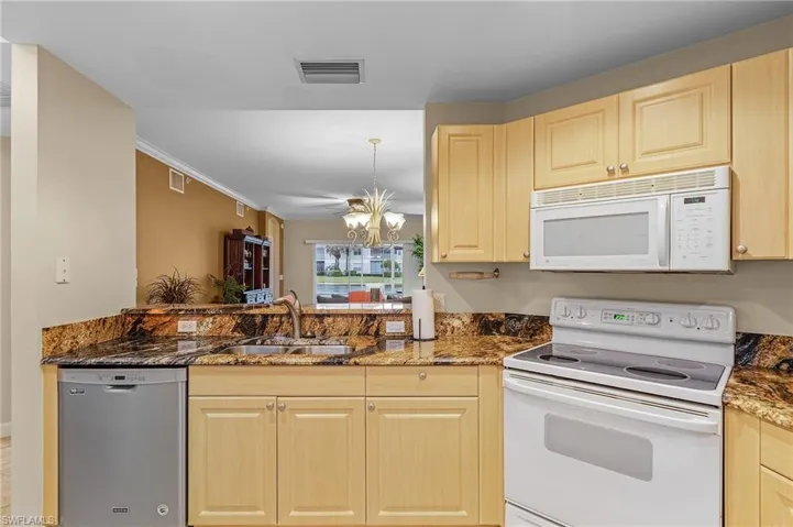 Kitchen with dark stone counters, a notable chandelier, white appliances, light brown cabinetry, and sink