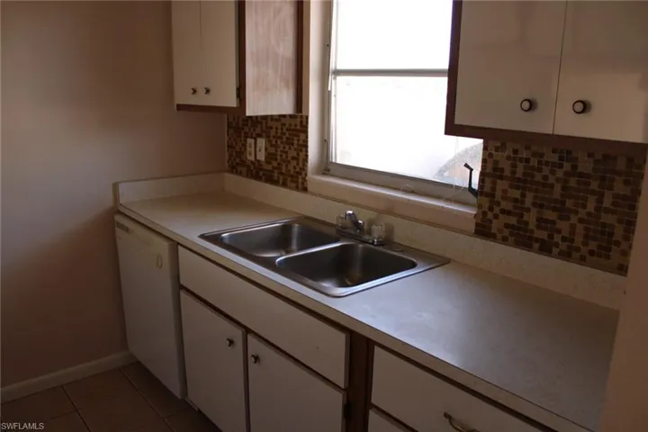 Kitchen with backsplash, white dishwasher, sink, light tile patterned floors, and white cabinetry