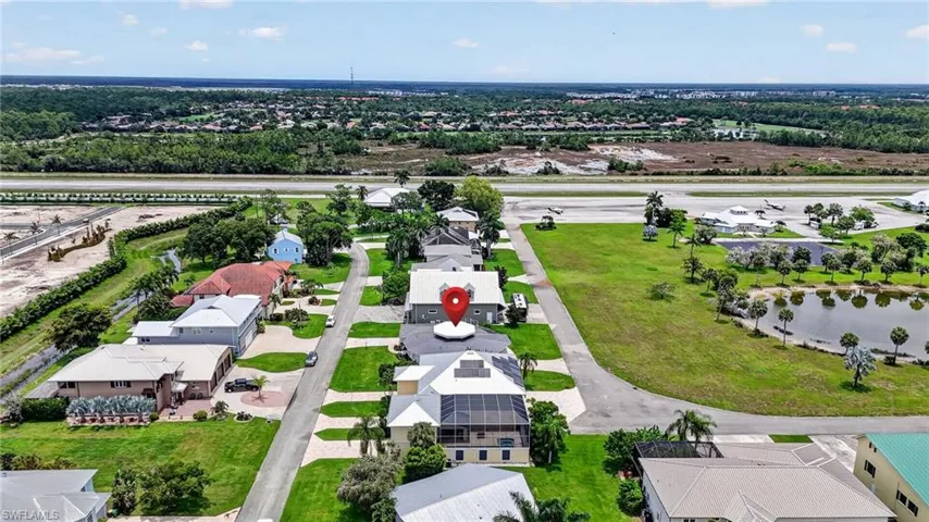 Aerial perspective showcasing a residential street with diverse architectural styles, surrounded by green spaces and distant tree lines and runway.