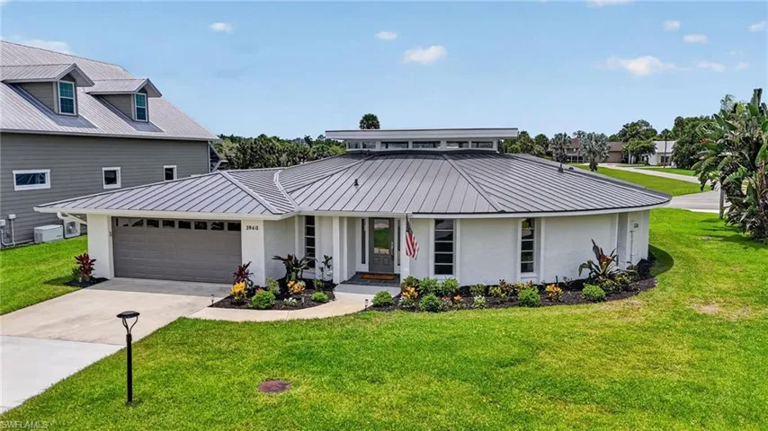 Modern circular architecture featuring a metal roof, white exterior, integrated garage, car/plane-port and a manicured lawn with mature landscaping