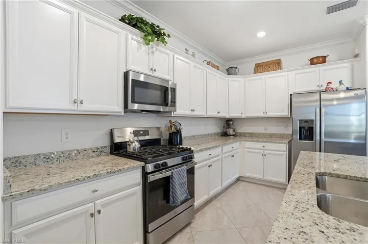 Kitchen with appliances with stainless steel finishes, white cabinetry, ornamental molding, light tile patterned flooring, and light stone counters