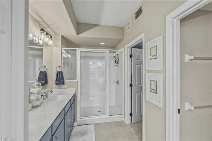 Bathroom featuring a dual-sink vanity with white countertops and dark cabinetry, a glass-enclosed shower, and neutral-toned tile flooring