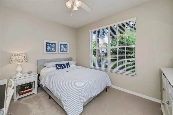 Bright bedroom featuring a large window with white blinds, neutral carpeting, and a ceiling fan with light kit