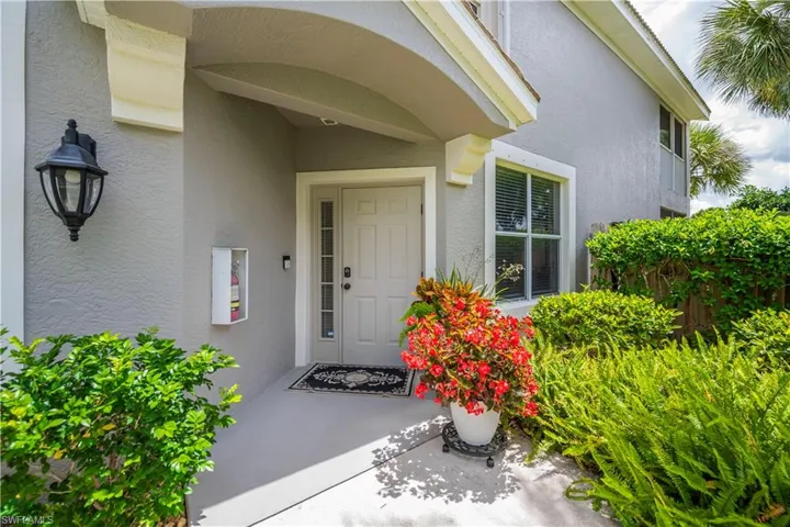 Inviting entryway featuring an arched portico, decorative exterior lighting, a light-colored entry door with sidelights, a large window with blinds, and mature landscaping