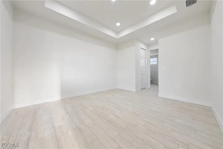 Spacious room featuring light-toned wood-finish flooring, white walls, a tray ceiling, recessed lighting, and a white paneled door