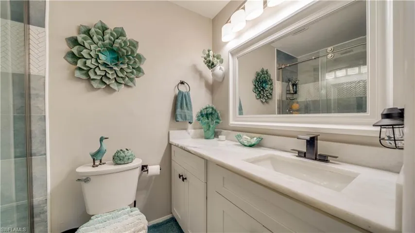 White shaker-style vanity with a solid surface countertop, integrated sink, and matte black faucet, featuring a large framed mirror with multi-bulb overhead lighting