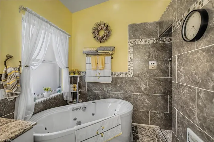 Oval jetted tub with tile surround, accent mosaic tiling, and a window with white sheer drapery