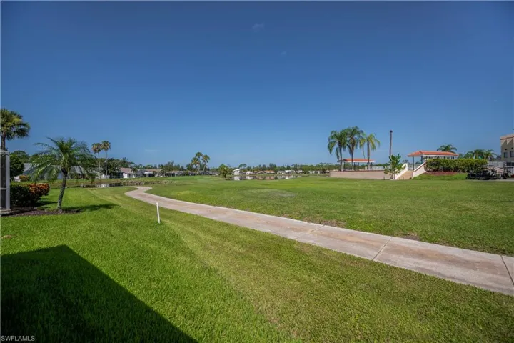 Expansive green space featuring a paved pathway, mature palm trees, distant water, and a covered pavilion structure