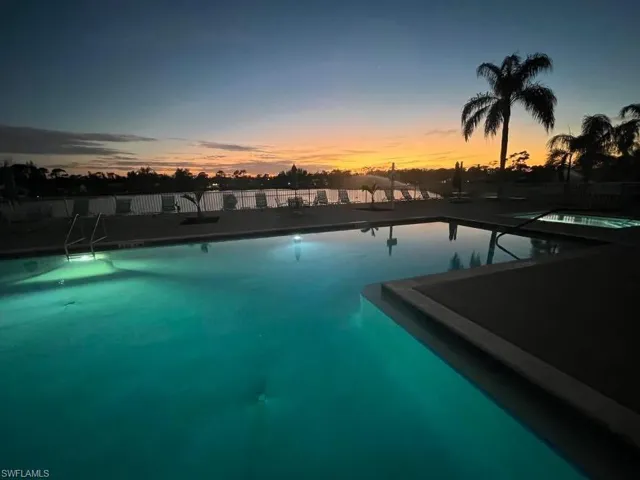 Illuminated swimming pool with an integrated spa, bordered by concrete decking and a metal fence