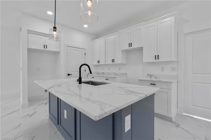 Kitchen with white cabinetry, light marble finish flooring, light stone countertops, a center island with sink, and hanging light fixtures