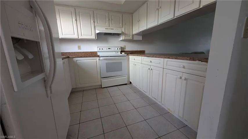 Kitchen with white appliances, white cabinets, light tile patterned floors, under cabinet range hood, and dark countertops