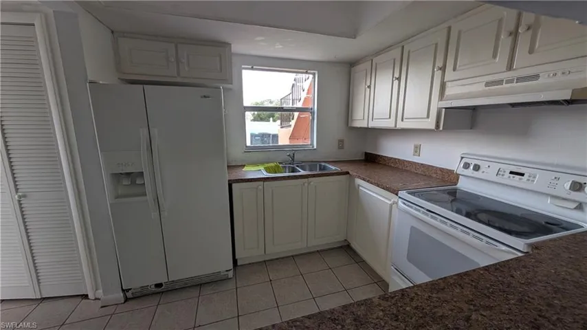 Kitchen featuring white appliances, dark countertops, under cabinet range hood, light tile patterned floors, and white cabinetry