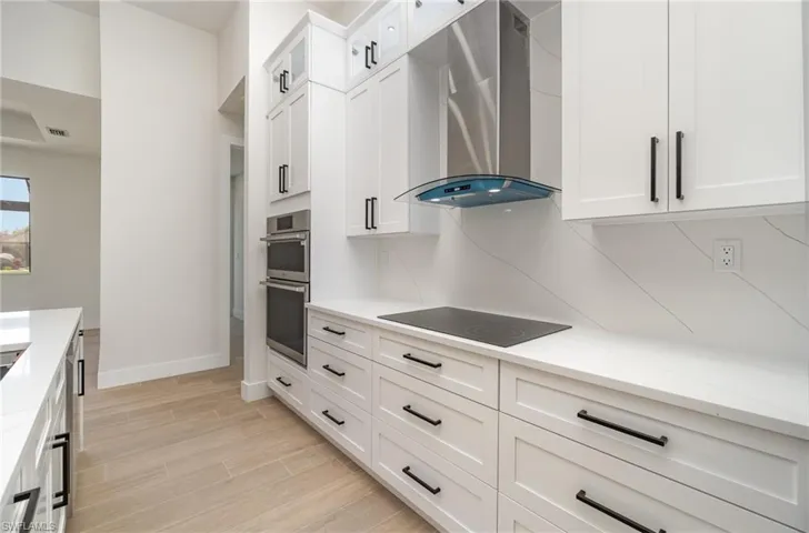 Kitchen with decorative backsplash, stainless steel double oven, white cabinetry, wall chimney exhaust hood, and black electric stovetop